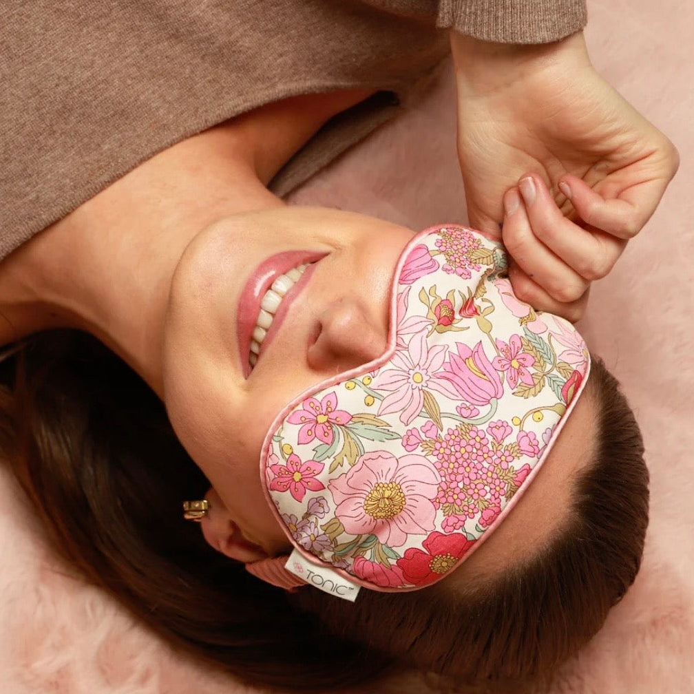 A woman lying down wearing a pink floral sleep mask, with her hand adjusting the strap. She is wearing a brown knit sweater against a soft pink background.