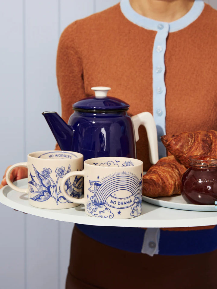 Person holding a tray with a blue teapot, mugs, and pastries.