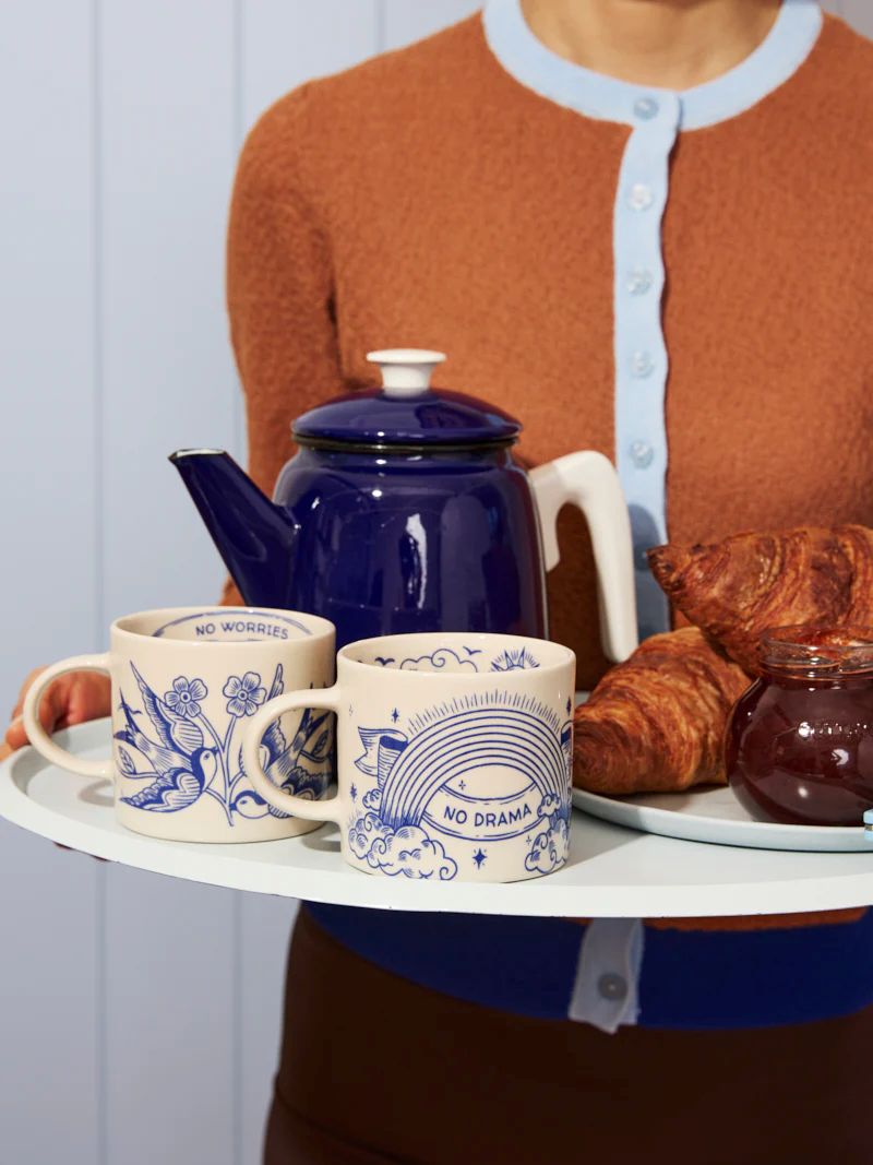 Person holding a tray with a blue teapot, mugs, and pastries.