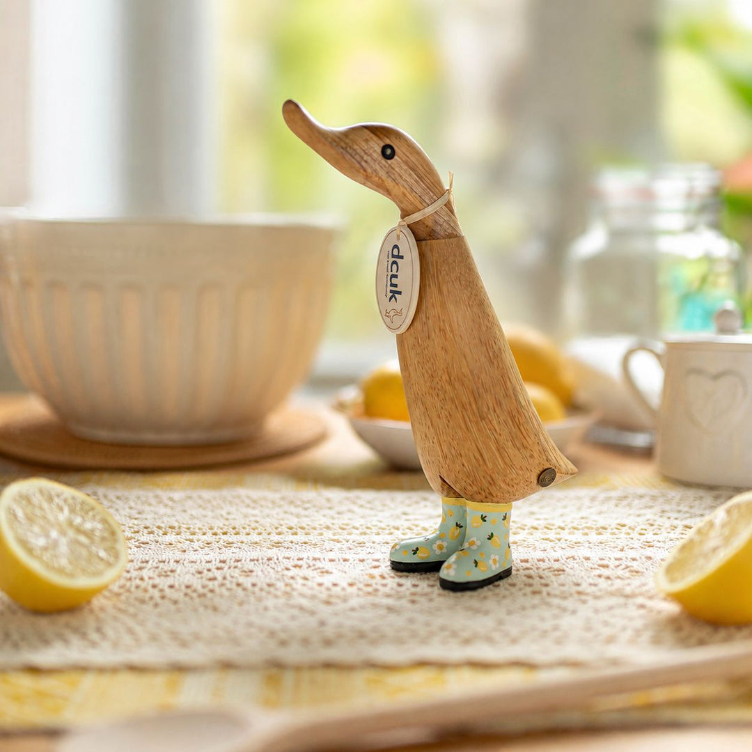 A wooden duck-shaped kitchen utensil with a light natural wood finish and pastel blue rain boots with yellow polka dots, standing on a table runner with lemons and ceramic bowls in a softly blurred kitchen setting.