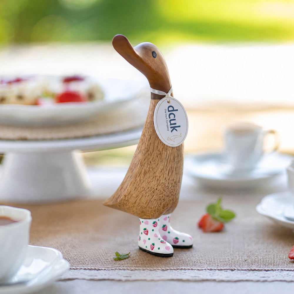 A wooden duck figurine with a natural wood finish and white rain boots printed with pink strawberries, standing on a table runner with a blurred breakfast setting in the background. A hanging tag reads 'dck'.