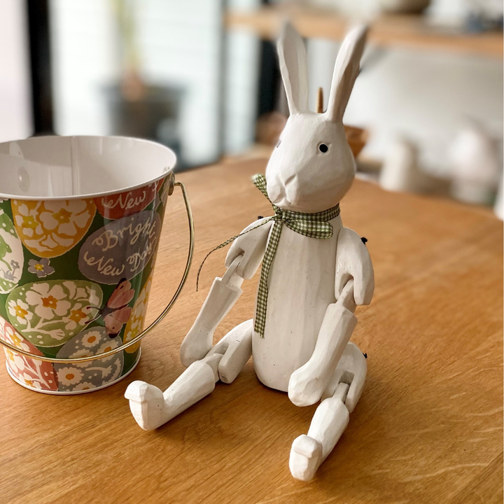White rabbit figurine with a green bow tie on a wooden surface next to a floral-patterned bucket.