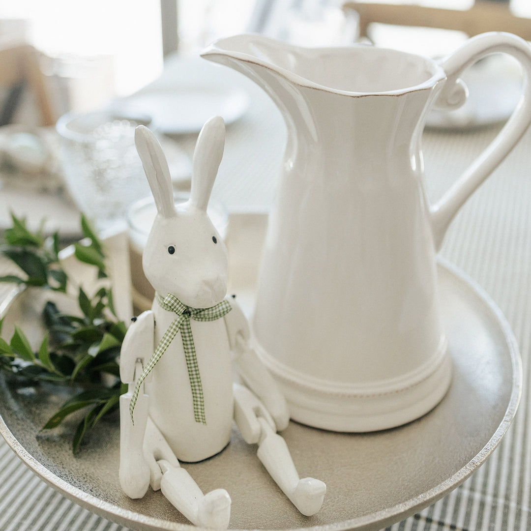 White wooden rabbit figurine with a green ribbon next to a white pitcher on a table.